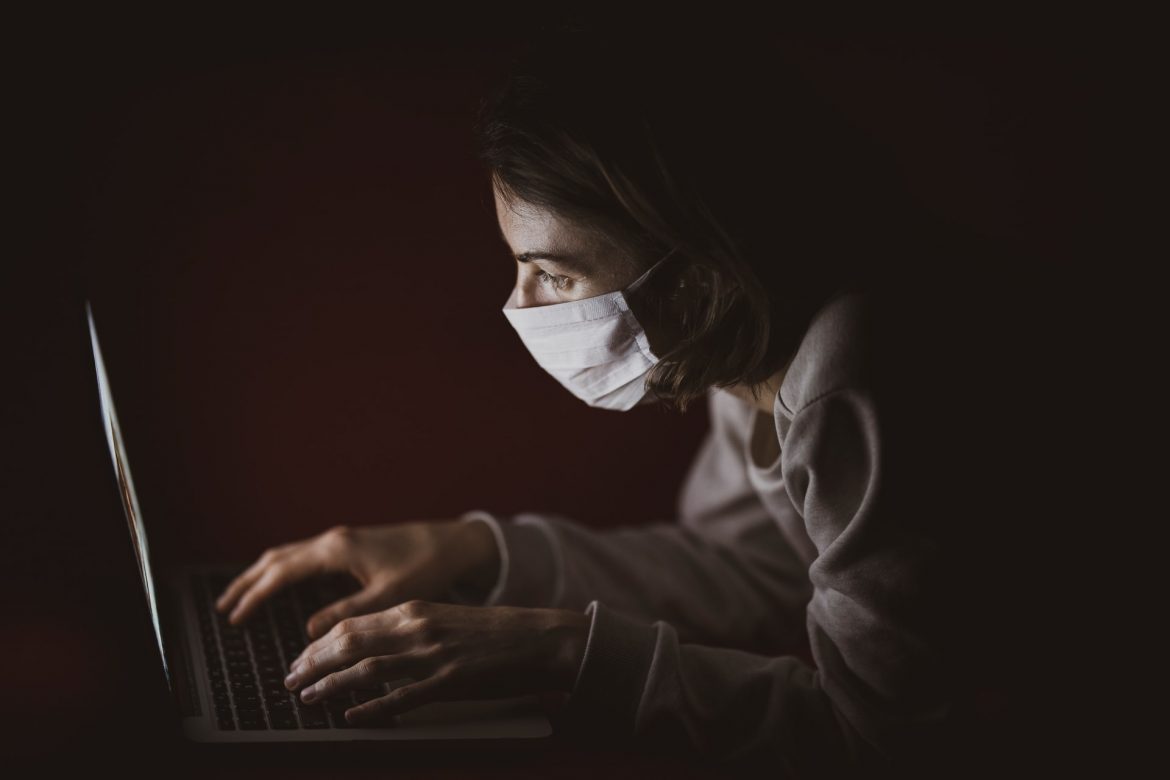 A woman wearing a face mask and staring at her laptop seated in a dark room